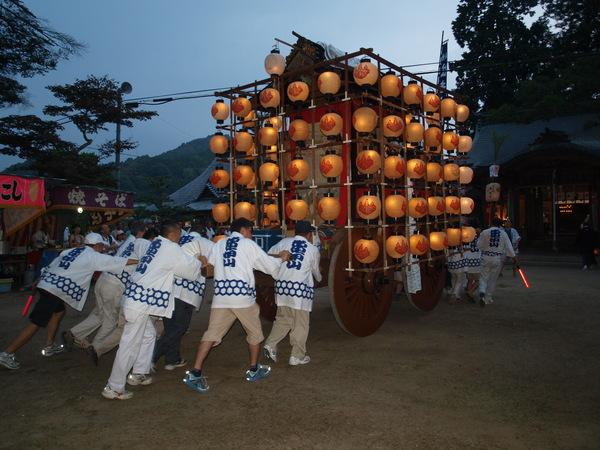 白色の法被を着た男性陣が提灯を灯した山車を一生懸命押している写真